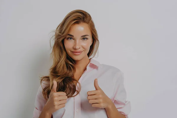 Pretty young European woman with satisfied expression recommends product or best service keeps two thumbs up gives praise wears shirt looks self confident at camera poses against grey studio wall