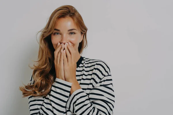 Beautiful cheerful woman giggles positively covers mouth with hands tries to hide emotions reacts on something funny dressed in striped jumper isolated over grey background blank space away.