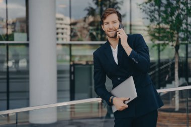 Kendine güvenen genç sakallı iş adamı iş görüşmesi yapıyor, cam binanın dışında duruyor ve iş arkadaşlarıyla cep telefonunda konuşurken rahat bir poz veriyor.