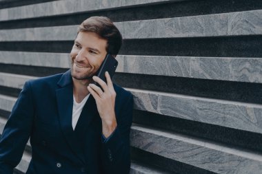 Mola sırasında cep telefonuyla konuşan şık ceketli çekici genç bir ofis çalışanı. İş merkezinin önünde dururken aile içi güzel konuşmalar yaparken samimi bir şekilde gülümsüyor.