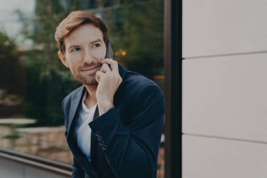 İş iletişimi. Genç yakışıklı şehir iş adamı cep telefonuyla müşteriyi dinliyor, akıllı telefondan bir şeyler tartışıyor, yansıtıcı pencereli binanın yanında tek başına duruyor.