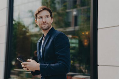 İşverenine proje detayları hakkında kirli sakallı mesajlar atan başarılı yakışıklı iş adamı, elinde akıllı telefon tutarken büyük bir pencereyle şehir manzarasını yansıtıyor.