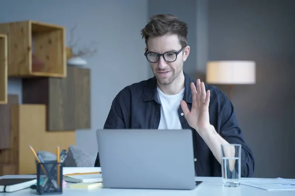 Young handsome austrian man freelancer in glasses waving hand in hello gesture during video call on laptop computer, male employee greeting colleagues at online meeting while working remotely at home