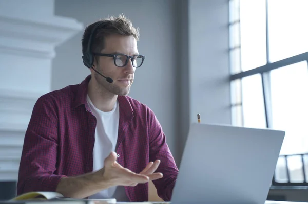 European man online tutor wearing glasses sitting at desk in headset and talking by video call on laptop computer while working remotely from home. Distance education and e-learning concept