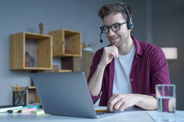 Smiling German guy in casual clothes sitting at home in wireless headset and taking part in online meeting with colleagues, male freelancer using laptop computer during video call with client