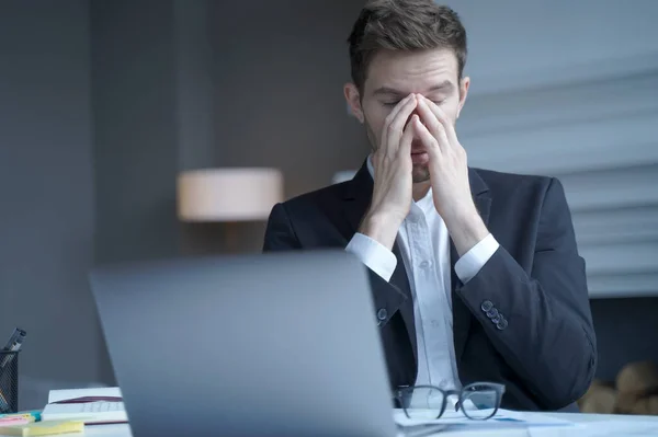Overworked male entrepreneur sits at home office while took off glasses massages bridge of his nose with eyes closed, exhausted man suffering from heavy headache and fatigue. Health problem at work
