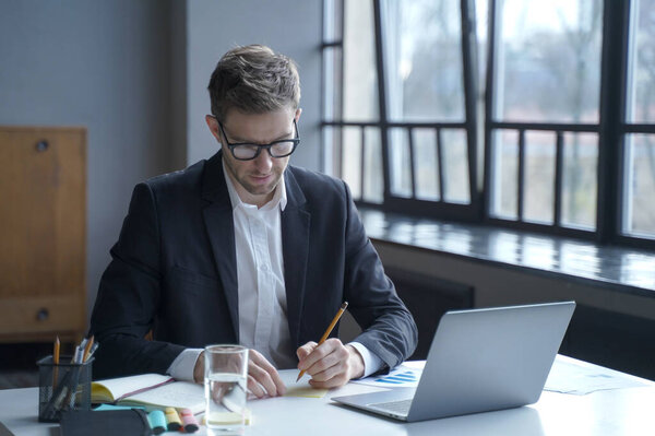 Immersed in work. Focused young German male banker writes down memos on sticky notes to remember important details to share for upcoming online meeting while sitting at desk at modern home office