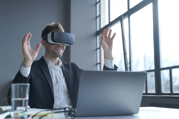 Employee experience with virtual reality. Young businessman in suit sitting at office table and using vr glasses goggles at work, male entrepreneur testing innovative method for business