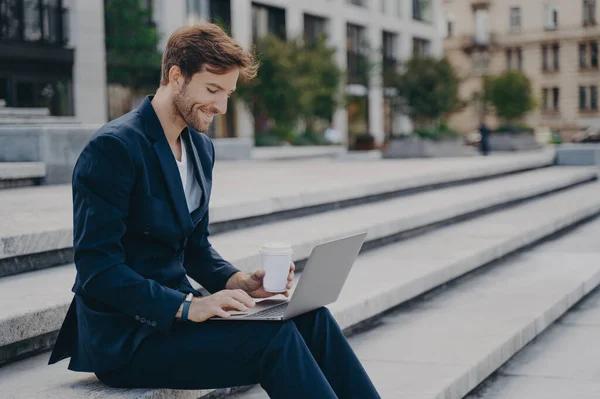 Young satisfied businessman in stylish dark blue suit sitting on stairs near office center outside with laptop and coffee to go in sunny summer day, working remotely online while having lunch break