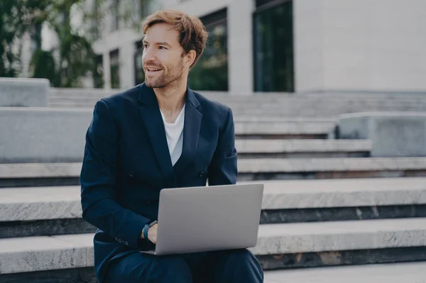 Smiling brown-haired neat unshaven office worker in stylish suit sitting outside on stairs in city center with laptop on his lap, checking important emails and reports from work. Remote job outdoors