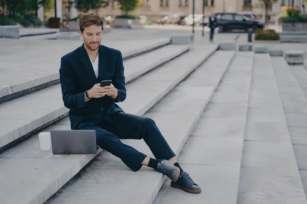 Young confident urban business professional man in stylish suit using smartphone while sitting on steps outside with laptop. Businessman holding mobile phone using app, typing messages or urgent email