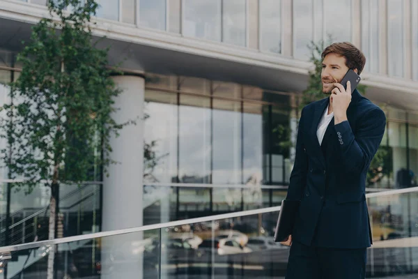 Sideways shot of handsome bearded man calls via smartphone discusses successful project after presentation dressed in black formal suit poses near office business building outdoor. Job and technology