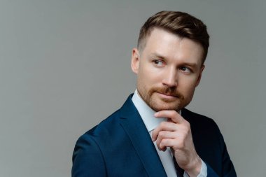 Thoughtful bearded businessman holds chin considers something looks away makes decision dressed in formal clothes looks forward future opportunity isolated over grey background blank space for text