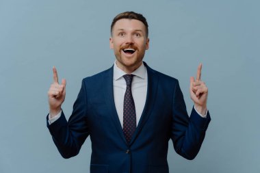 Positive optimistic businessman points above demonstrates something with glad expression dressed in formal suit white shirt and tie poses against blue background shows top advertisement deal on market