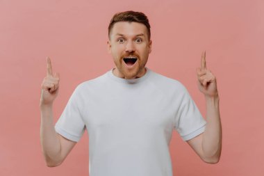 Half length shot of impressed bearded European guy points above with both index fingers gasps fromm wonder has surprised face expression wears basic white t shirt isolated over pink background