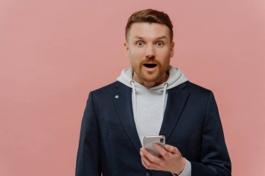 Şok olmuş bir yüz ifadesiyle kameraya bakarken akıllı telefon tutan, şaşkın, kızıl saçlı adam pastel pembe arka plan üzerinde izole edilmiş yakışıklı adam. İnsan duyguları kavramı