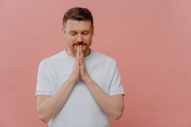 Believing in something good. Young handsome man in white t shirt keeping hands in praying gesture and eyes closed, asking for good luck or making wish while standing isolated over pink background