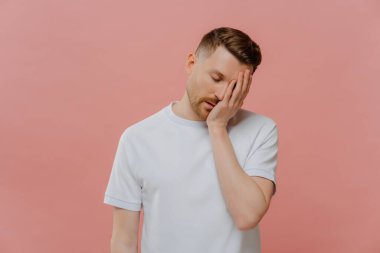 Genç, beyaz sakallı, gözleri kapalı, uykulu hisseden, pastel gül arka planında poz veren, elini yüzüne koyan ve hayal kırıklığı gösteren genç bir adam.