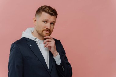 Young bearded man touching chin and thinking about something or having doubts while posing on pink background. Serious businessman with thoughtful face expression making decision, copy space for text