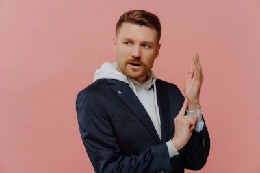 Sakallı, genç, kızıl saçlı bir adam nabzını ölçüp yan bakıyor. Havalı giyinmiş, parmaklarını bileğinde tutan, pastel pembe arka planda soyutlanmış bir adam.