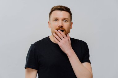 Stüdyoda, siyah tişörtlü, ağzı eliyle kaplayan ve kameraya şaşkınlıkla bakarken gri arka planda şoke olan, şaşırmış ve şaşırmış yakışıklı bir kızıl adam görüntüsü var.