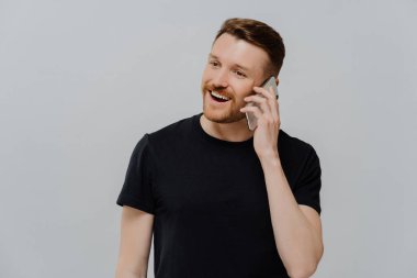 Kirli sakallı mutlu genç adam cep telefonuyla konuşuyor ve tatmin oluyor, olumlu haberler alıyor, kızıl saçlı adam yan bakıyor ve gri stüdyo duvarına karşı gülümsüyor.