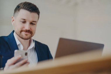 Genç, sakallı, kulaklık takan genç bir iş adamı dizüstü bilgisayarın önünde otururken internetten toplantı yapıyor şık takım elbiseli bir erkek yönetici ofis masasının arkasında otururken cep telefonu kullanıyor.