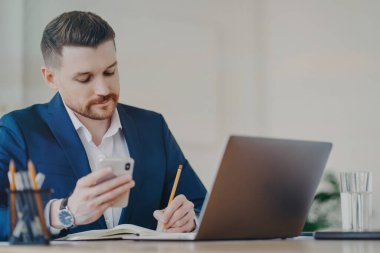 Elinde cep telefonu olan genç ve başarılı bir işadamı ofiste dizüstü bilgisayarın önünde otururken önemli verileri not defterine yazıyor, koyu mavi takım elbise ve pahalı bir saat takıyor.