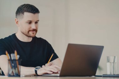 Yakışıklı sakallı genç serbest çalışan ya da sıradan çalışan elinde kalemle Webinar seyreden ya da internette sörf yapan evden uzakta modern bir dizüstü bilgisayar üzerinde çalışan biri.