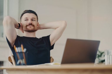 İş yerinde rahat rahat giyinen mutlu, genç bir ofis çalışanı kollarını başı bağlı, yan bakıyor ve gülümsüyor, dizüstü bilgisayarla masada oturuyor. Serbest çalışan mutlu görünüyor çünkü iş bitti.