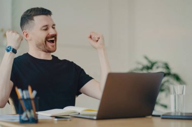 Excited young freelancer or businessman showing winning or lucky gesture demonstrating his happiness and celebrating successful deal during video call, sitting at table in office or home. Business concept