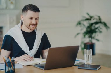 Attractive successful male freelancer wearing earphones taking part in online meeting while sitting in front of laptop at his cozy workplace at home or office, manager in casual wear sitting behind office desk