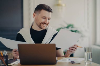 Kablosuz kulaklıklarda başarılı bir iş adamı, belgeleri analiz ediyor ve evden çalışırken iş sonuçlarından memnun görünüyor. Erkek ofis çalışanı işyerinde oturup kendini mutlu hissediyor.