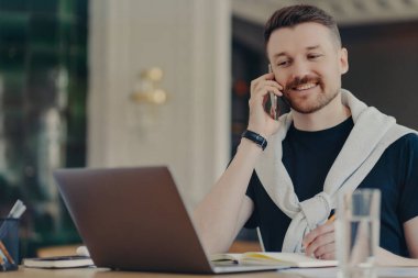 Genç başarılı girişimci ya da erkek serbest çalışan cep telefonuyla konuşan ve iş yerinde ya da evinde otururken dizüstü bilgisayara bakan, gülümseyen ve iş fikirlerini tartışan biri.