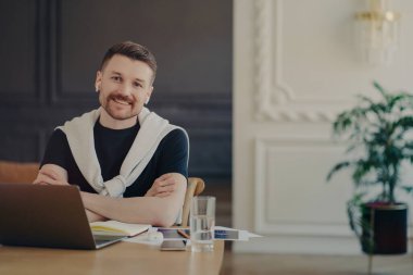 Genç erkek serbest çalışan, gülümseyen ve kameraya bakan, iş yerindeki bilgisayarının önünde oturan, siyah t-shirt giyen ve omuzlarının üzerinde beyaz süveter giyen.