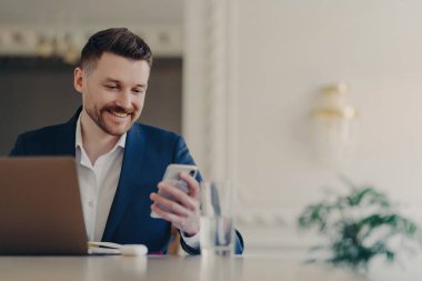 Successful handsome executive manager in formal suit looking at smartphone and smiling while reading messages or good news, sitting at his work desk in front of laptop in stylish office