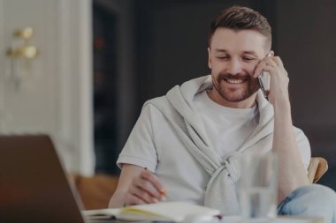 Attractive smiling male freelancer or businessman talking on phone while sitting at his work desk in front of laptop at home office making notes, wearing tshirt and sweater over his shoulders