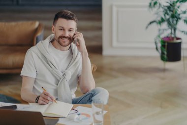 Attractive happy man freelance worker talking on mobile phone at spacious and stylish home office, making notes with pencil while casually sitting at his work desk wearing comfortable clothes