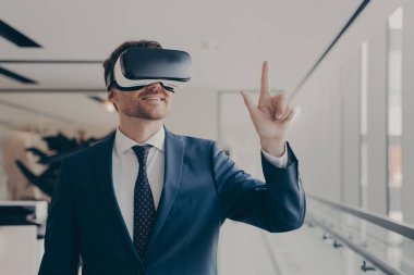 Cheerful businessman dressed formally using vr headset at work, wearing virtual reality glasses and working in augmented reality, pointing at virtual objects with finger. Futuristic technology in business