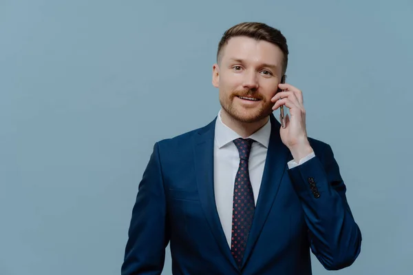 Making business call. Smiling businessman in suit talking on mobile phone with client or partner while standing isolated over light steel blue background, male entrepreneur enjoying conversation