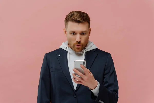 It can not be true. Impressed handsome redhead guy with beard in jacket over hoodie folding lips in wow, reading incredible news in his mobile phone while standing isolated over pink wall.