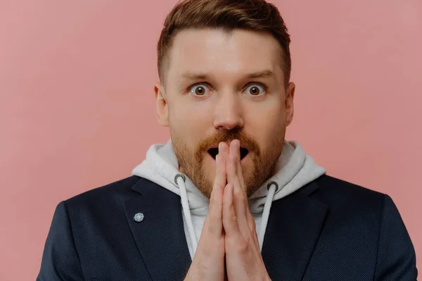Emotional bearded European man stares surprised clasps palms together stares bugged eyes gasps from wonder wears hoodie black jacket isolated over pink background has eyes full of disbelief.