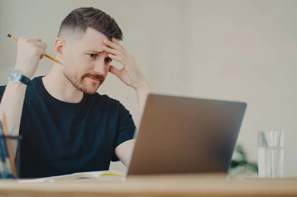 Worried depressed businessman or office worker holding pencil and nervously looking at laptop display, stressing about work results, having doubts about his business decision at workplace