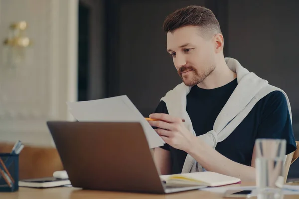 Concentrated man reads information or data from paper documents works on freelance poses at desktop against cozy interior dressed in casual clothes busy working. Male entrepreneur at coworking space