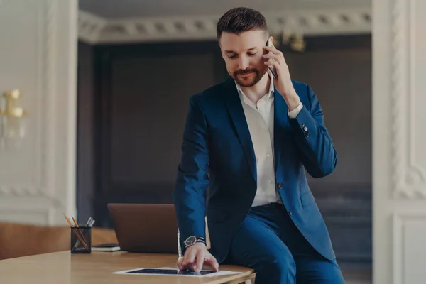Young handsome business professional in formal dark blue suit talking on phone and discussing project details presented on A4 document liying in front of businessman on work desk with laptop