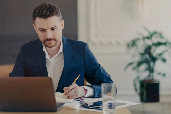 穿着正式深蓝色西服的成功的有吸引力的商人坐在办公室笔记本电脑前的办公桌前 在笔记本上写下了有关他的商业项目的重要数据 — 图库照片