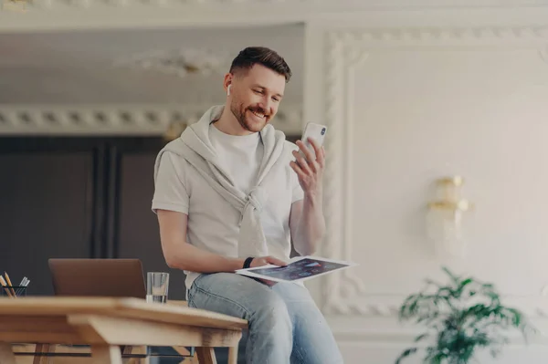 Smiling man freelancer wearing casual wear and earphones looking at phone and taking part in online meeting, casually sitting on desk and holding project documents in hand while working at home