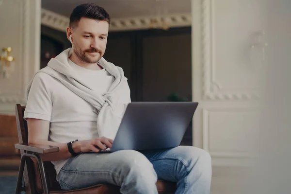 Young happy man freelancer in casual clothes wearing earphones using laptop while working from home, listening to music and looking at computer screen while sitting at modern home office interior