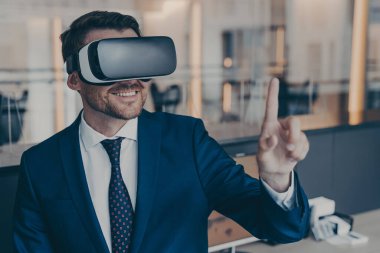 Resmi takım elbiseli gülümseyen genç sakallı finansörün yakından çekilmiş fotoğrafı sanal gerçeklikte VR gözlükleriyle vakit geçirmekten zevk alıyor ve işaret parmağıyla işaret ediyor. Siber uzay deneyim kavramı