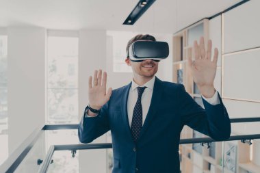 Takım elbiseli gülen ofis çalışanı VR kulaklık takıyor sanal gerçeklikte çalışırken havaya dokunuyor ve el kol hareketi yapıyor, iş adamı ofis koridorunda dururken 3D gözlükleriyle iş projesini yönetiyor.